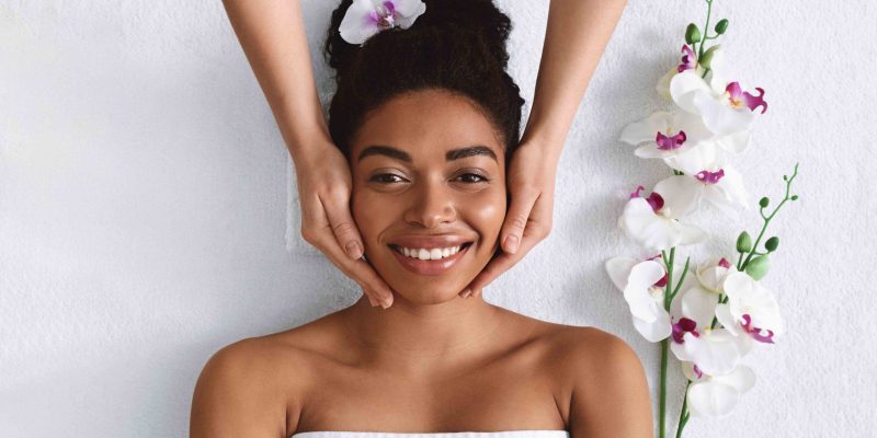 Aesthetic Procedure. Beautician making face massage for smiling black woman, top view